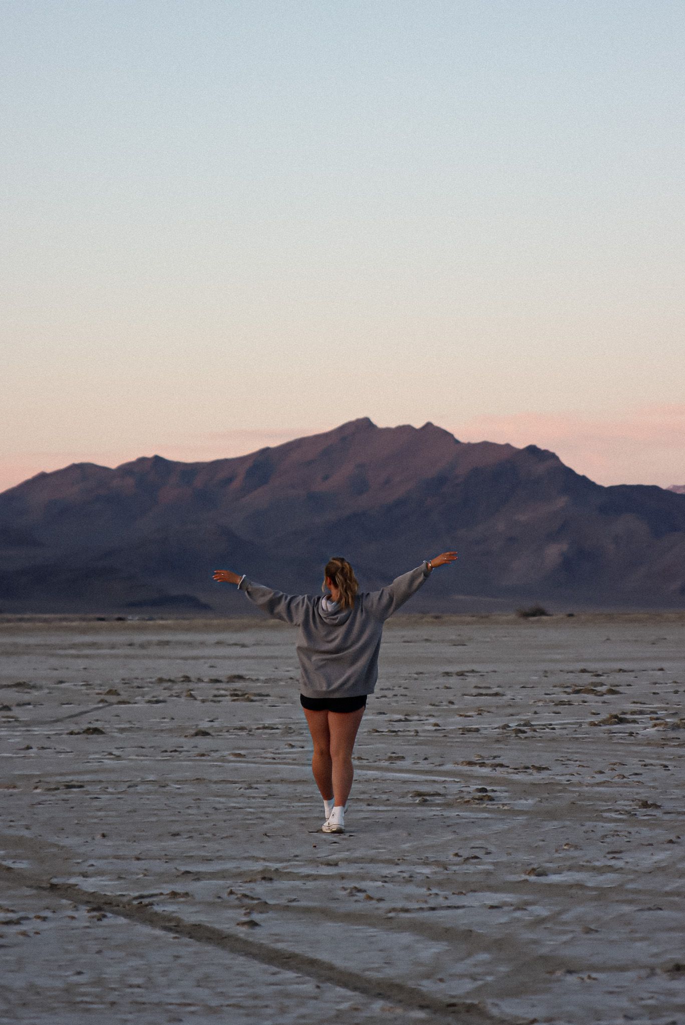 Cora Emilie Media A person stands with arms raised against a desert mountain landscape at sunset.