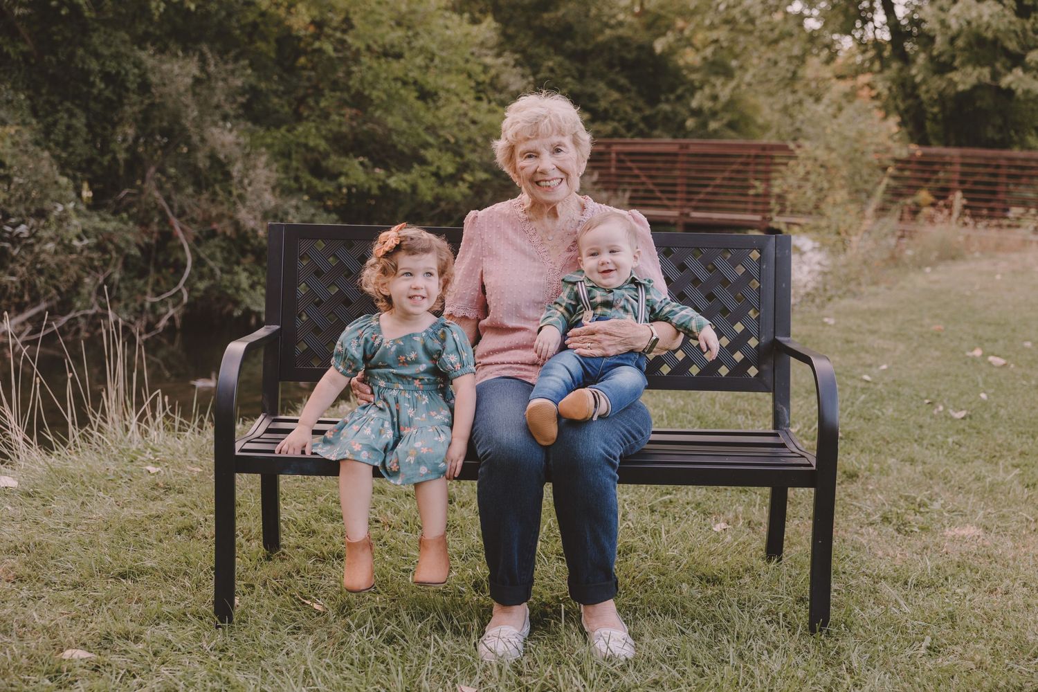A casual outdoor portrait shot of three generations sitting together on a black metal bench.