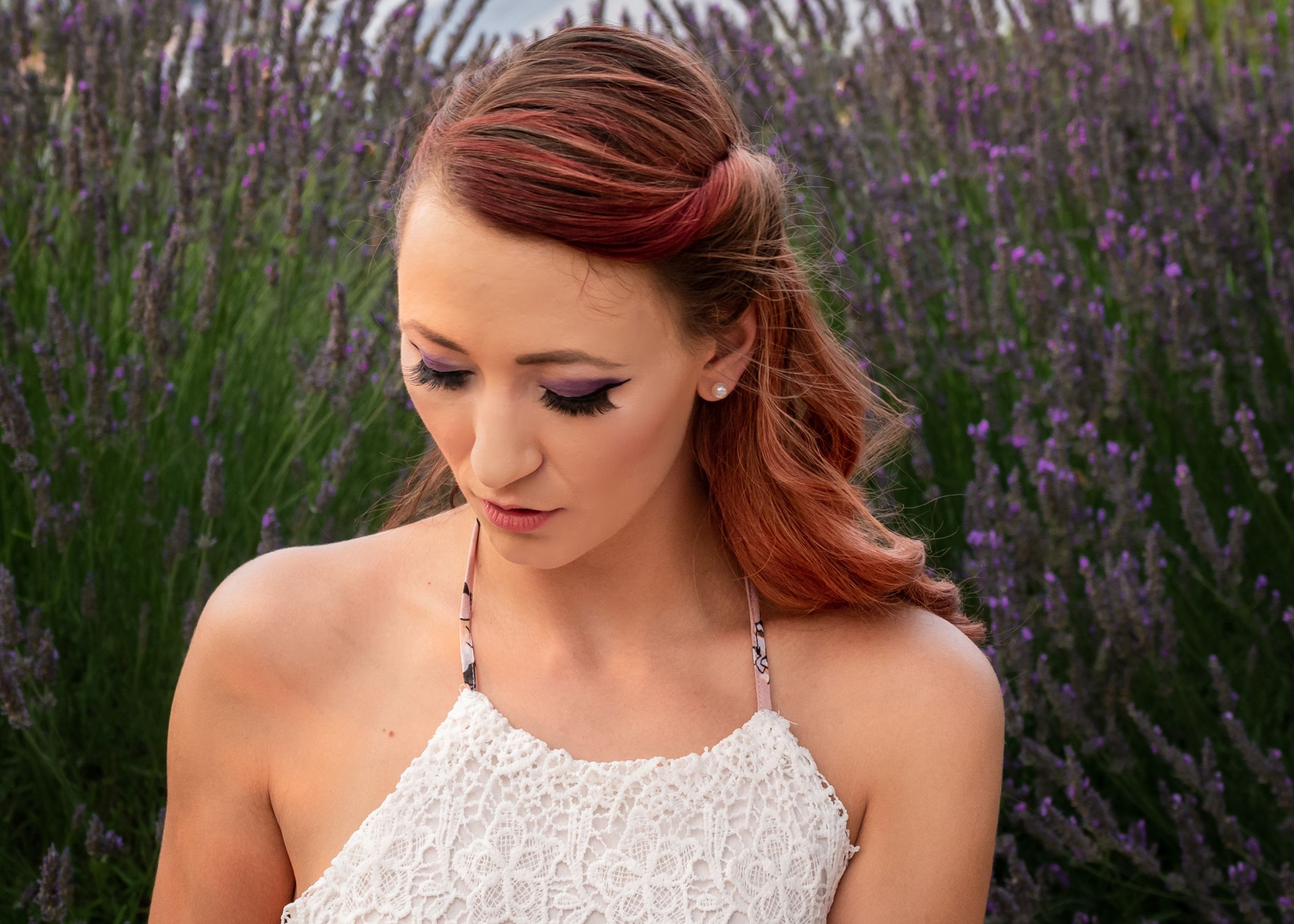 Young woman with auburn hair in white lace top gazes down among vibrant purple lavender flowers.