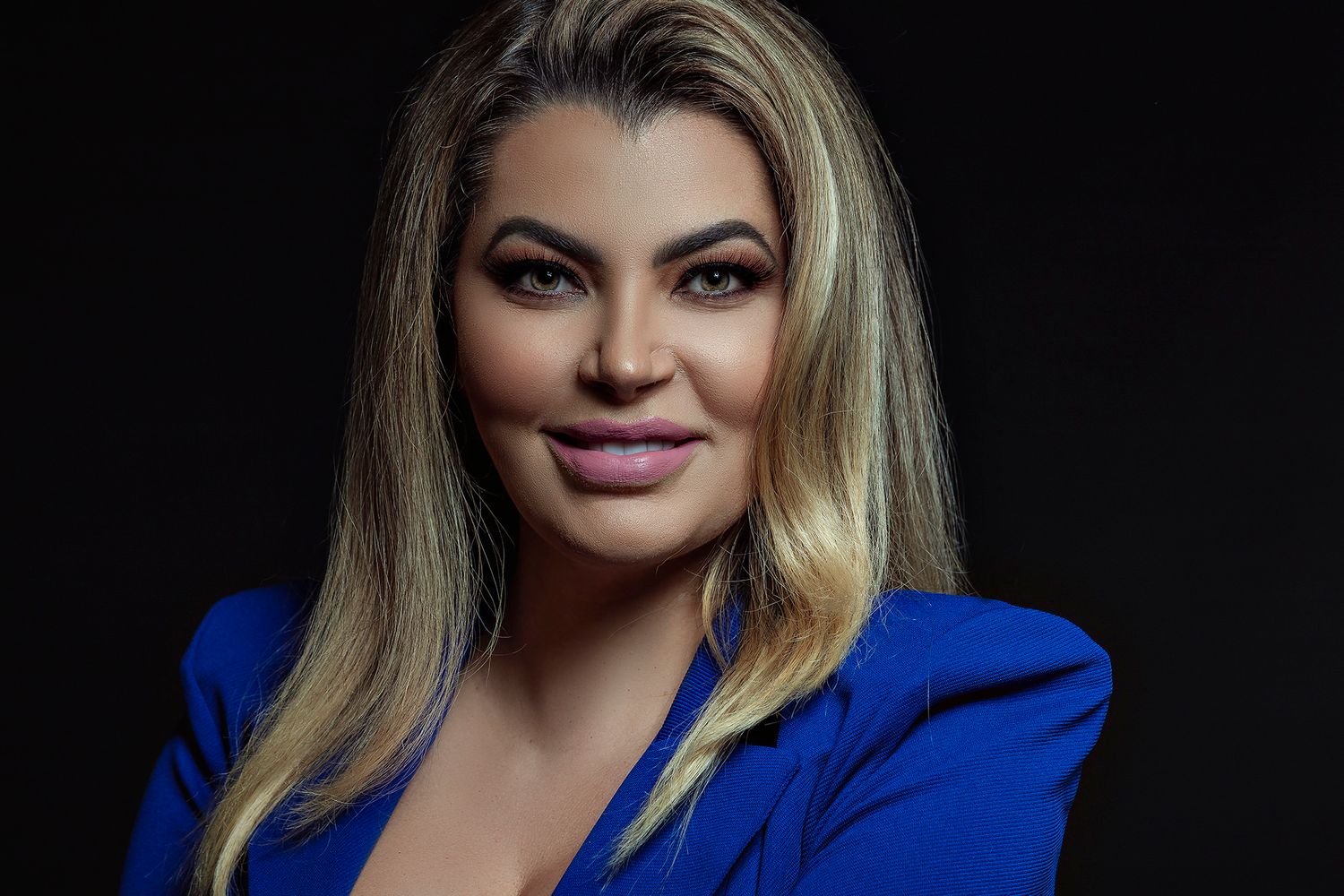 A professional headshot portrait of someone wearing a bright blue blazer against a dark background.