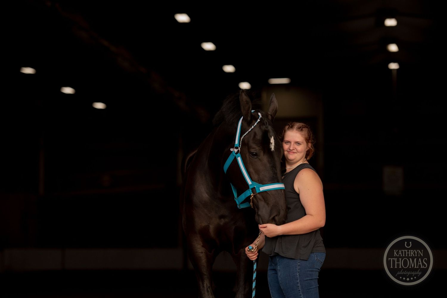 A beautiful horse with turquoise tack poses with its handler in a dark indoor arena setting.