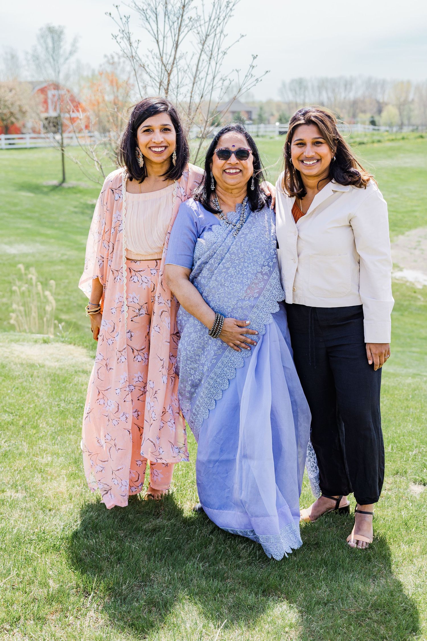 Three women pose together outdoors, wearing traditional and modern Indian clothing.