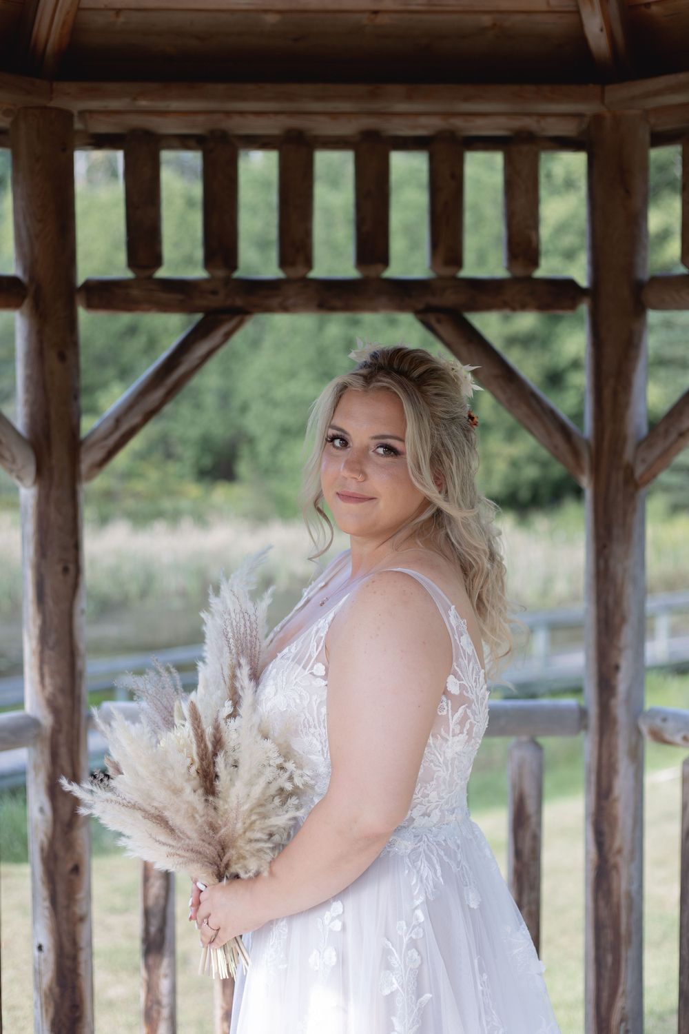 Bride in a lace wedding dress poses under a rustic wooden gazebo holding dried flowers.