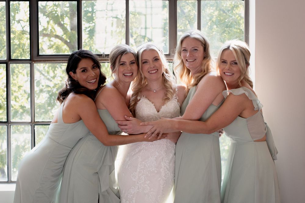Bride and bridesmaids in sage green dresses share a group hug moment at a wedding.