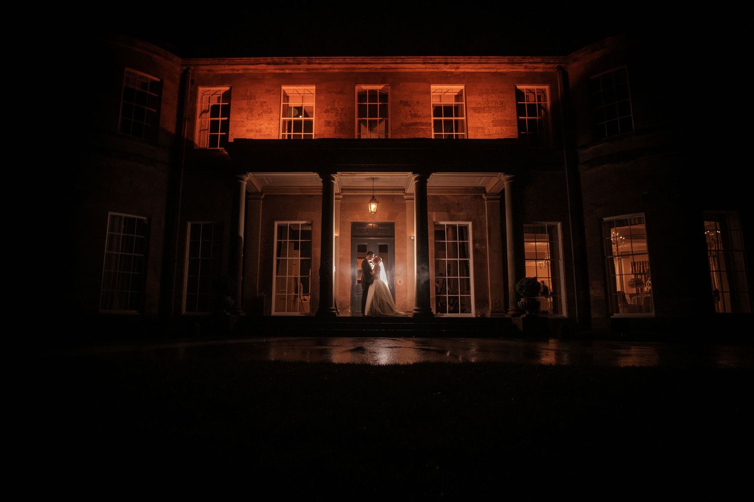A dramatic nighttime silhouette of a bride in a grand historic mansion doorway illuminated by warm lighting.