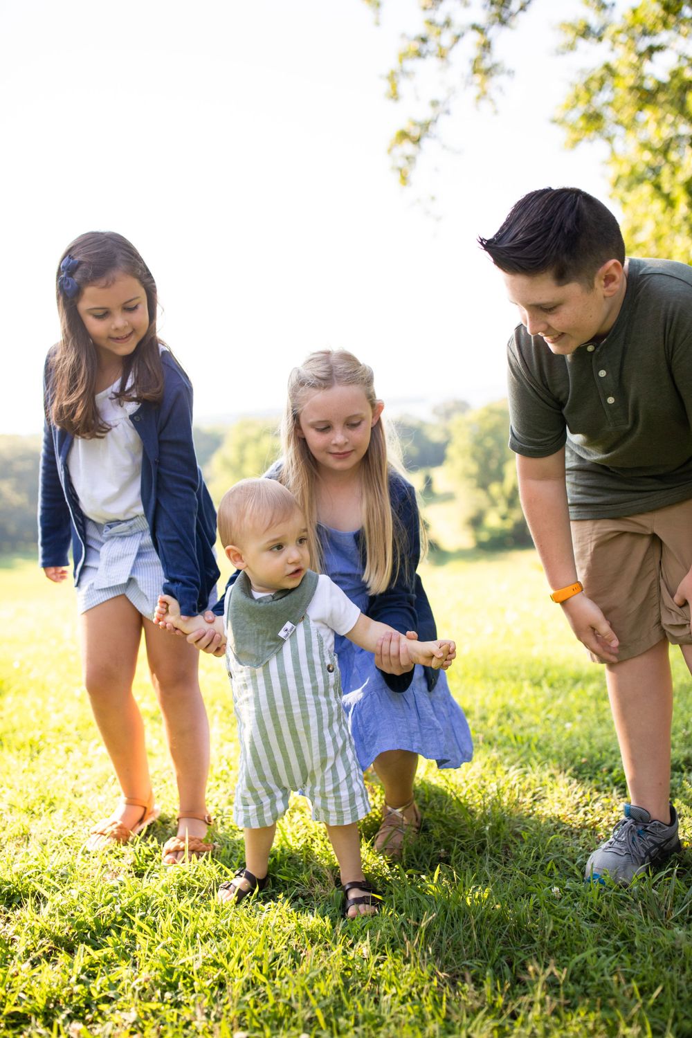 A group plays together in a sunlit grassy area while helping a toddler walk.