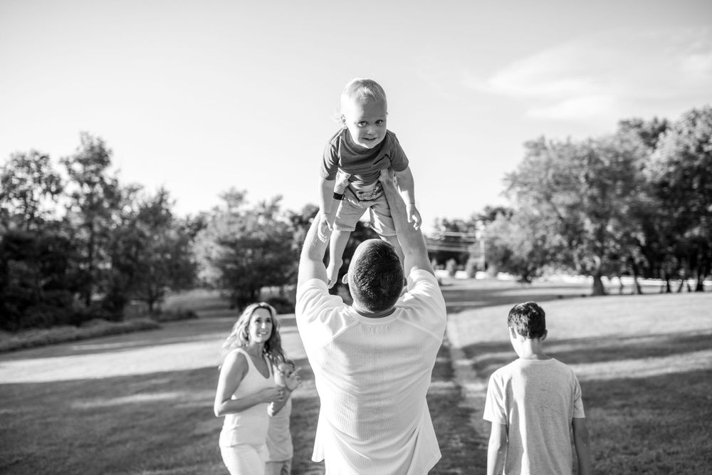 Black and white capture of family playing in park with child being lifted in air.
