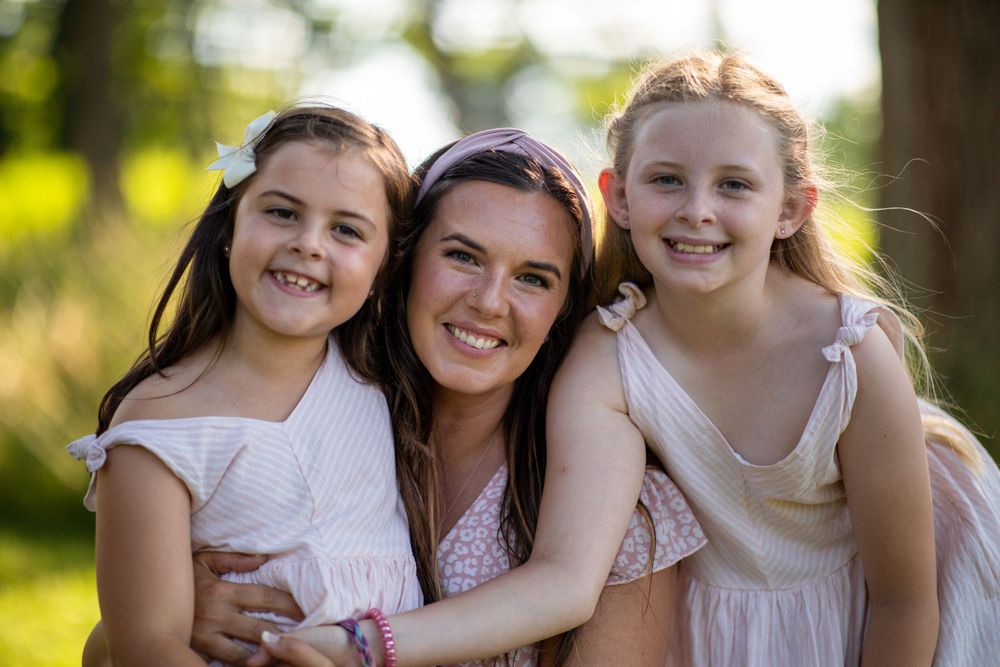 Three people pose together outdoors on a sunny day wearing light colored dresses.