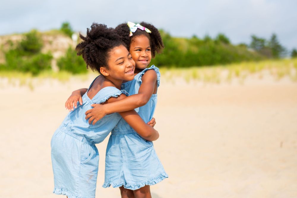Two children in matching blue dresses sharing a joyful embrace on a sunny beach.