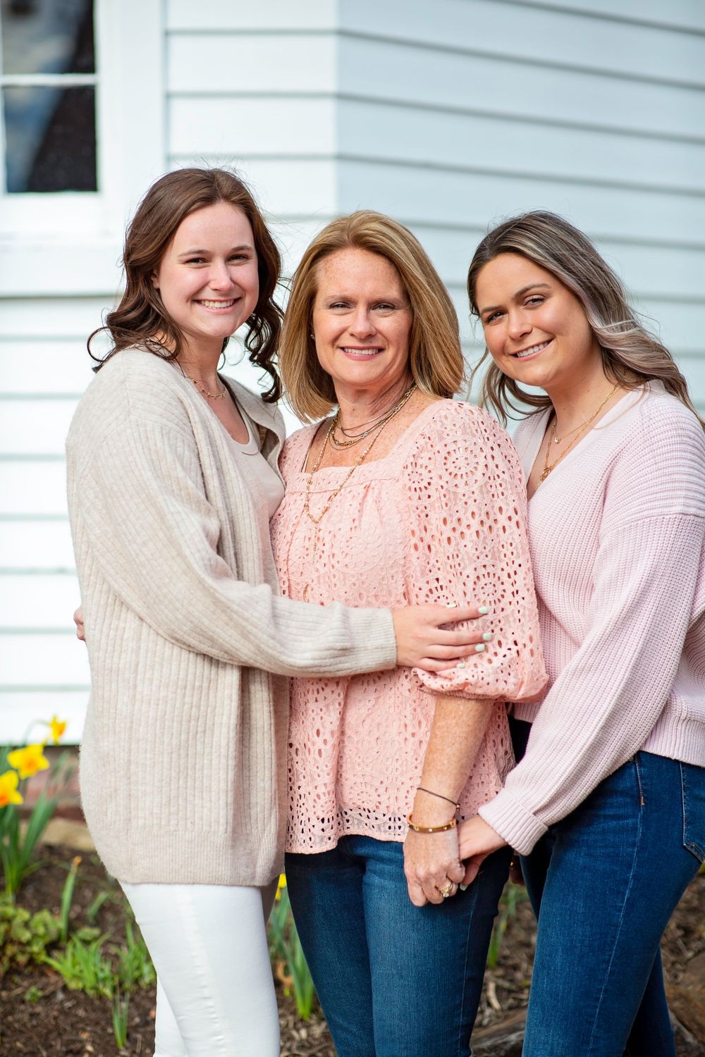 Three people embrace while wearing cozy sweaters in front of a white house with spring flowers.