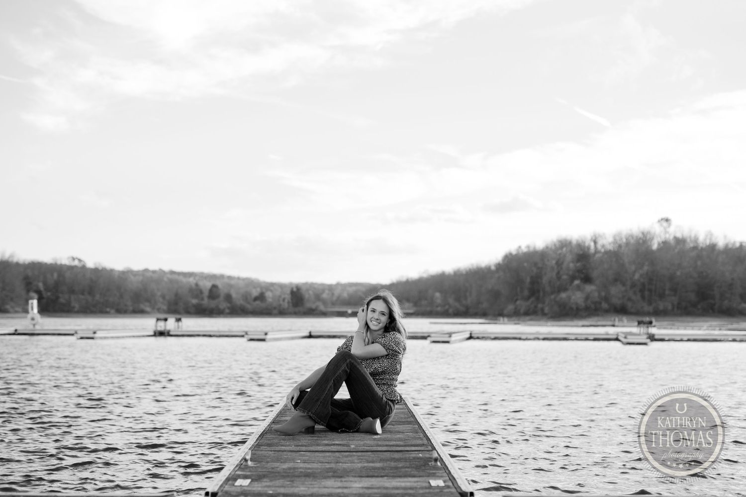 A black and white photo showing someone sitting on a wooden dock over a peaceful lake with trees in the background.