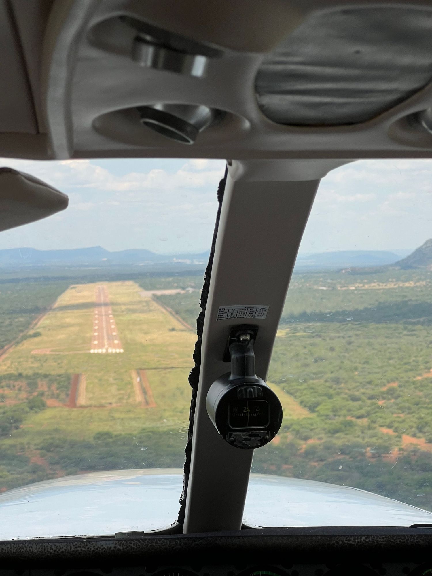 Aerial view from cockpit approaching a dirt runway surrounded by arid terrain and mountains.