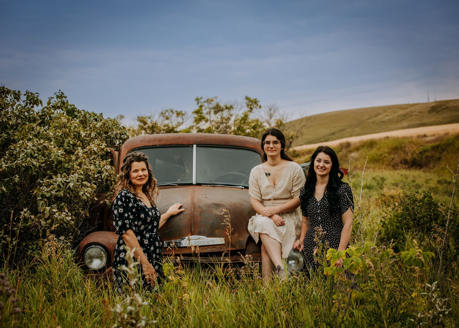 Three friends pose with vintage rusty car in grassy field with rolling hills.