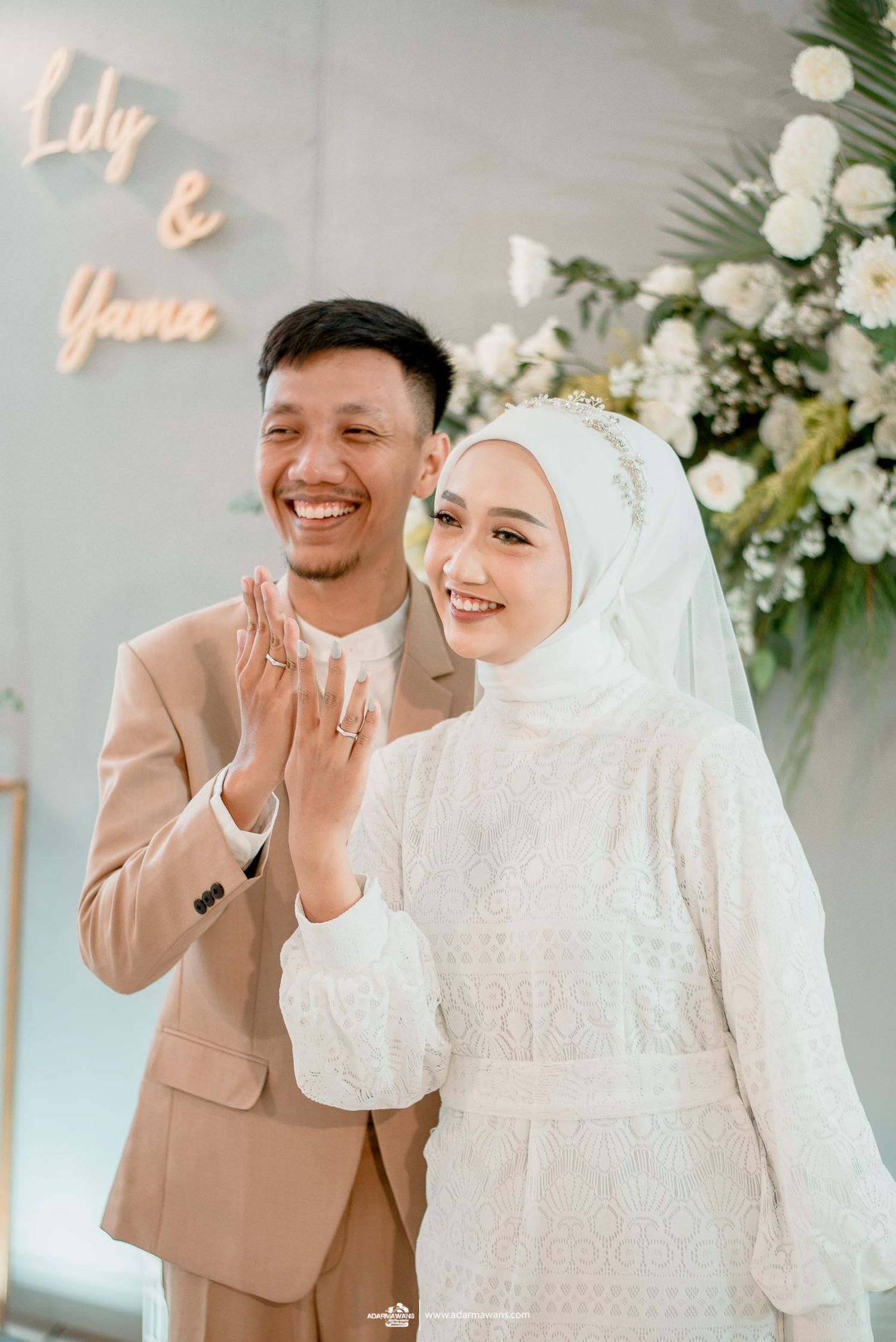 Happy newlyweds share joyful moment beneath neon sign and white floral decorations.