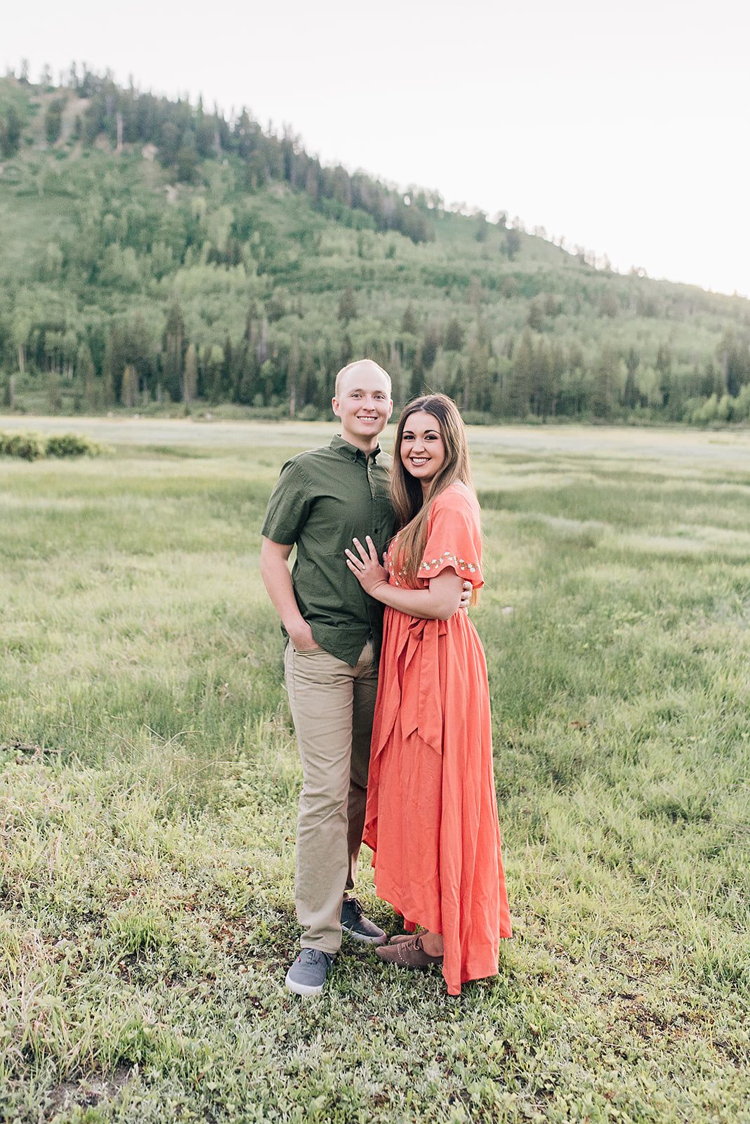 A couple embraces in a sunny meadow with forested hills in the background, wearing green and coral outfits.