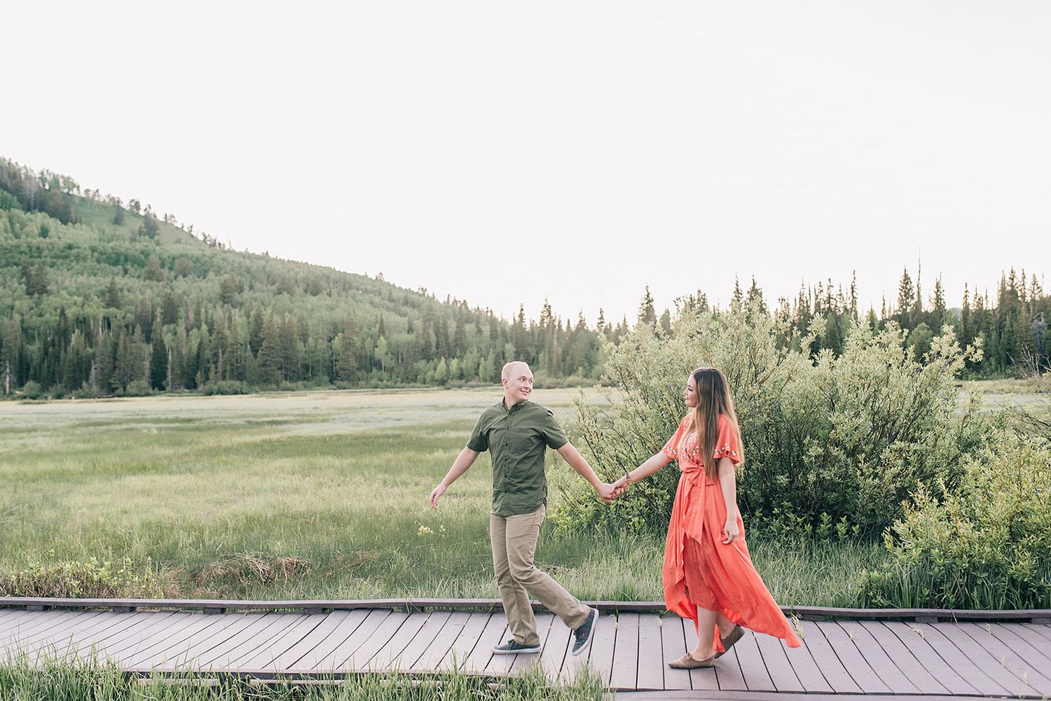 Couple balancing on wooden boardwalk surrounded by summer mountain scenery.