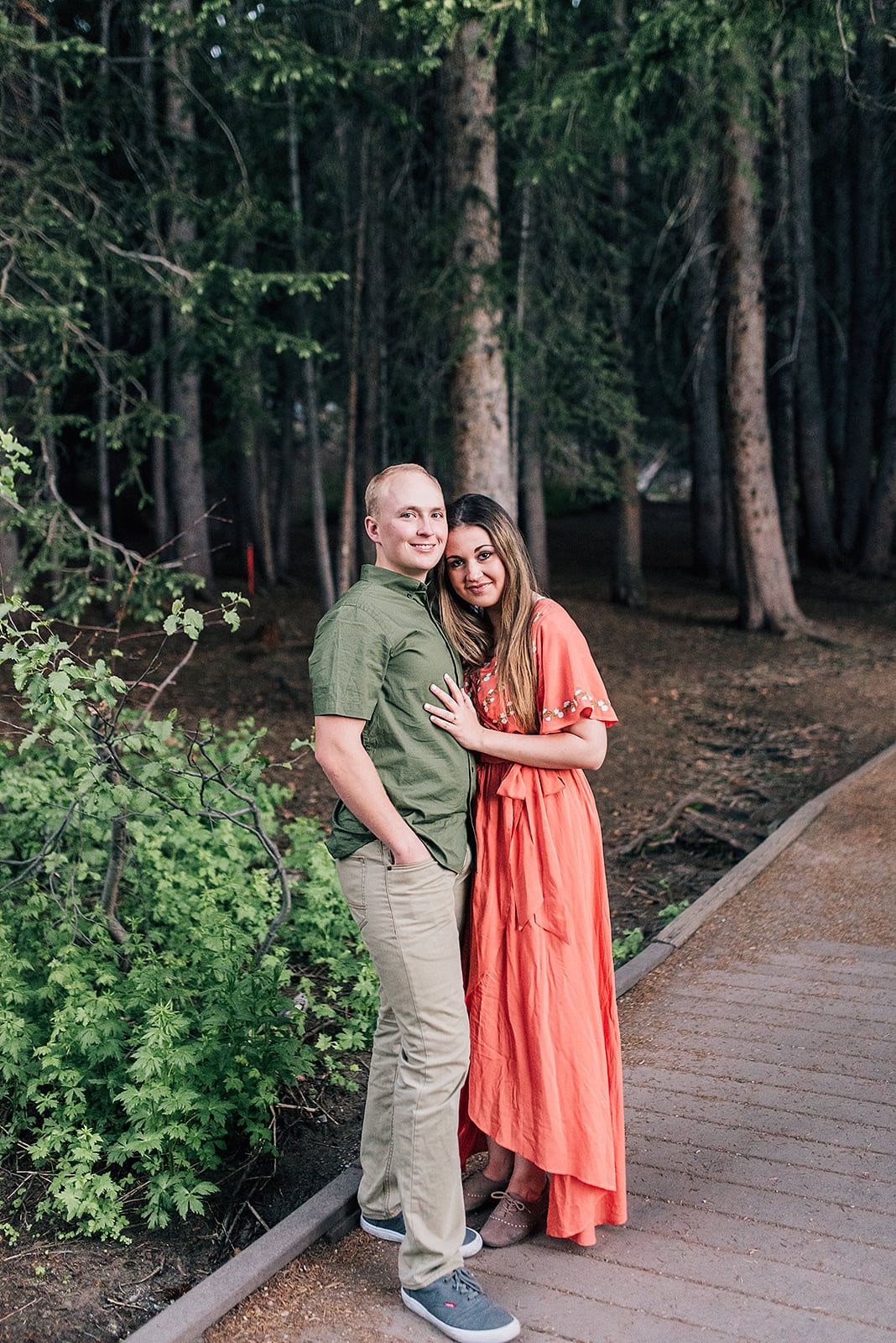 A couple poses on a wooden boardwalk surrounded by tall pine trees in matching green and coral outfits.