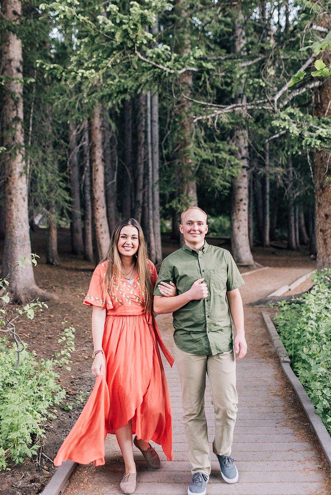 A couple walking hand in hand along a wooden pathway through a forest wearing coordinated outfits.
