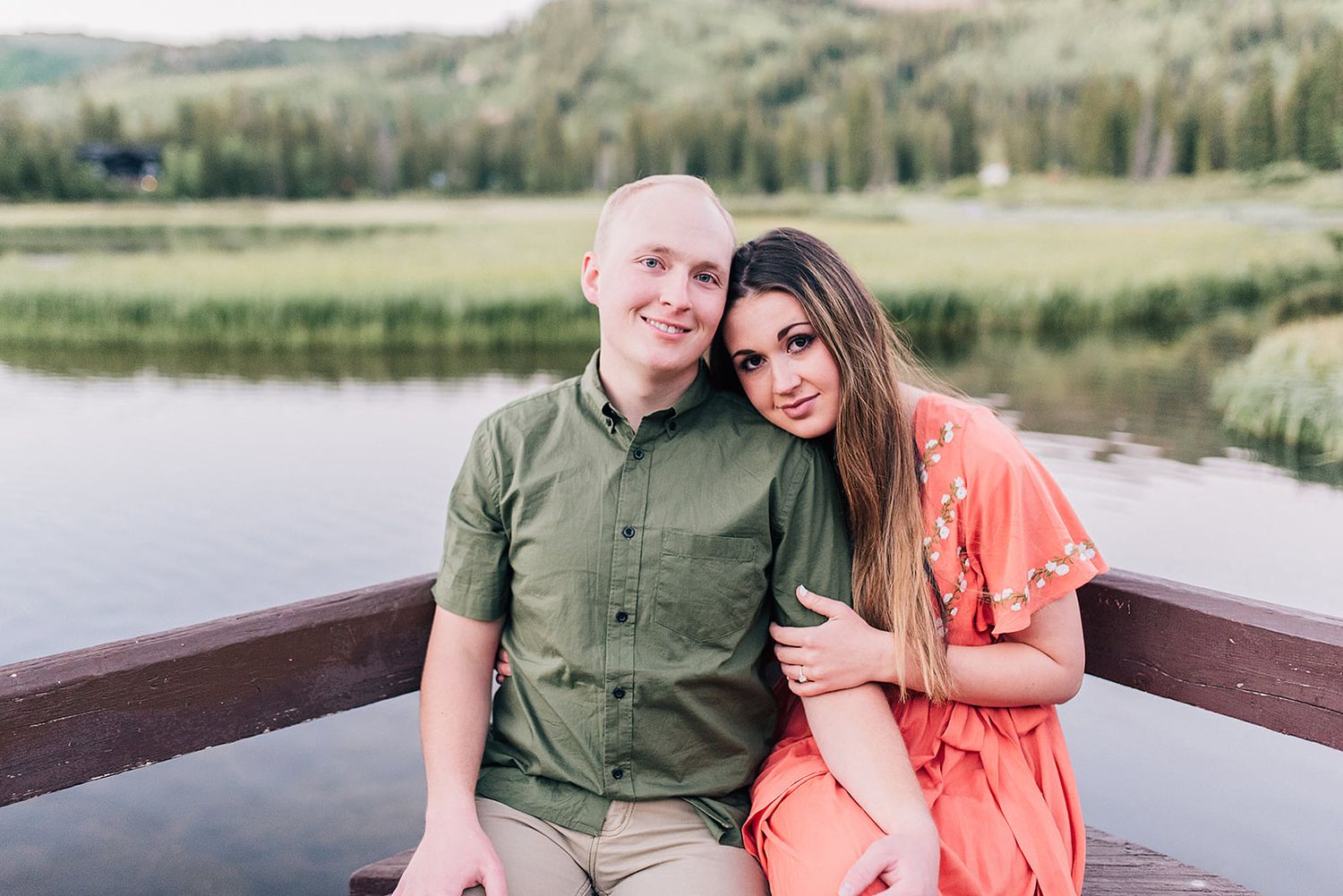 A couple sits together on a wooden dock overlooking a serene lake and mountain landscape.