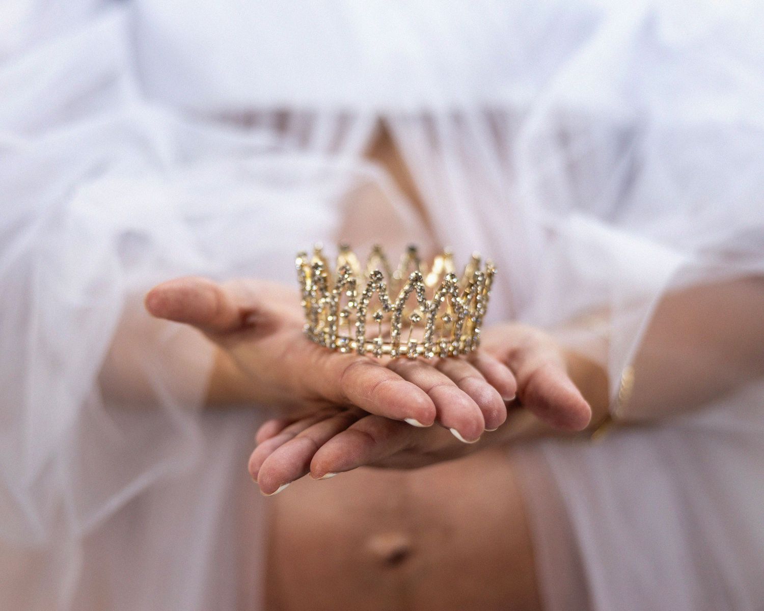 Delicate gold crown held in hands with white tulle fabric in dreamy setting.