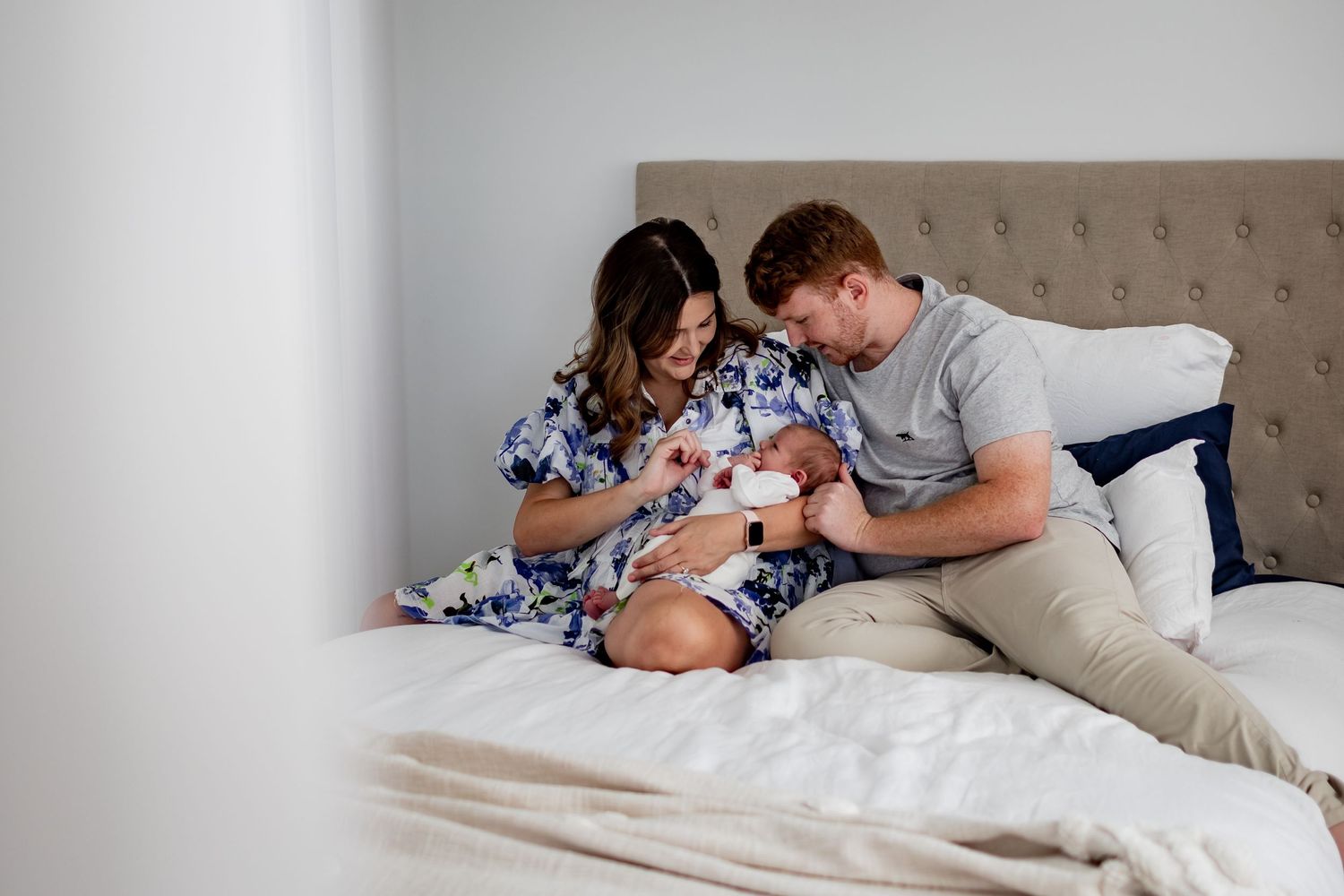 Family moment on bed with newborn baby in floral dress and grey attire.