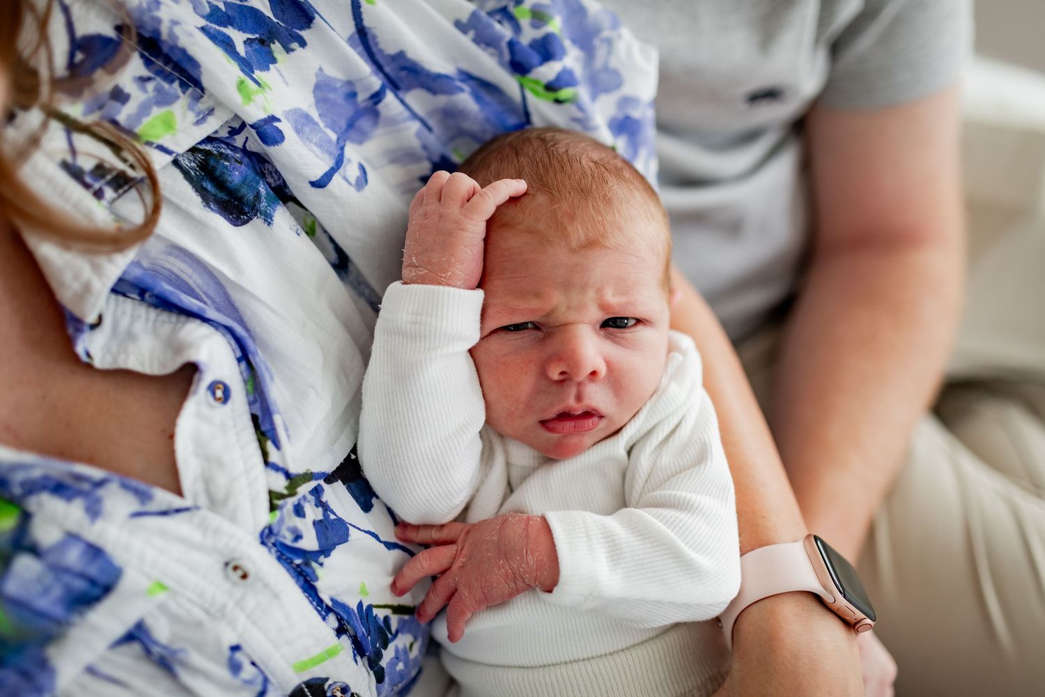 Newborn baby in white outfit touching their head while being held.