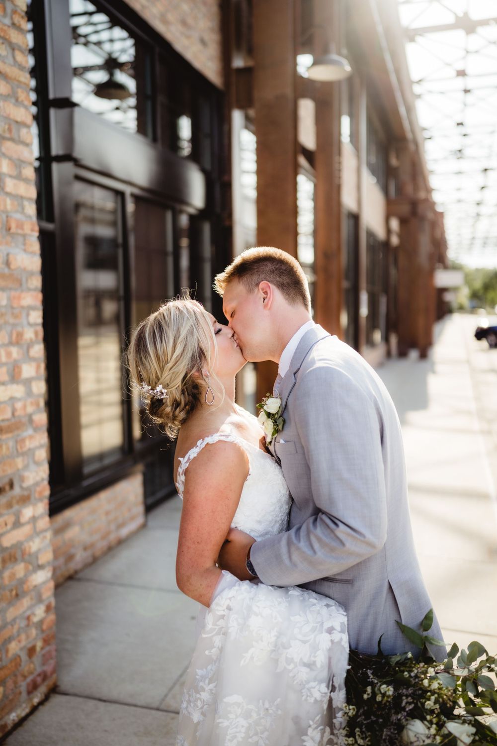 A romantic wedding couple sharing a kiss in front of a brick building with sunlit windows.