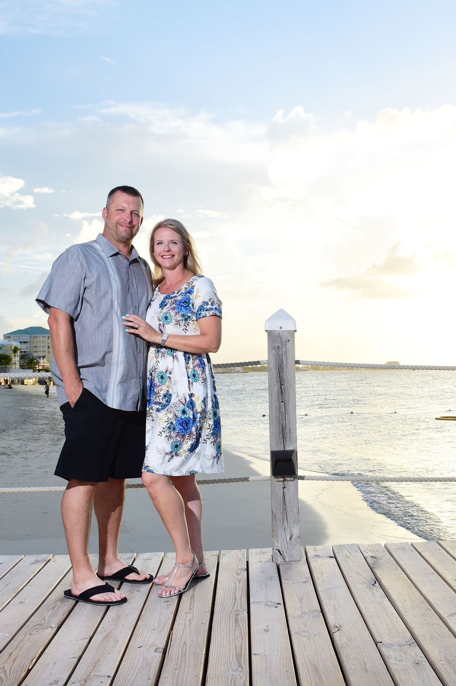 Couple stands on a wooden dock at sunset overlooking the ocean with a white post in the background.