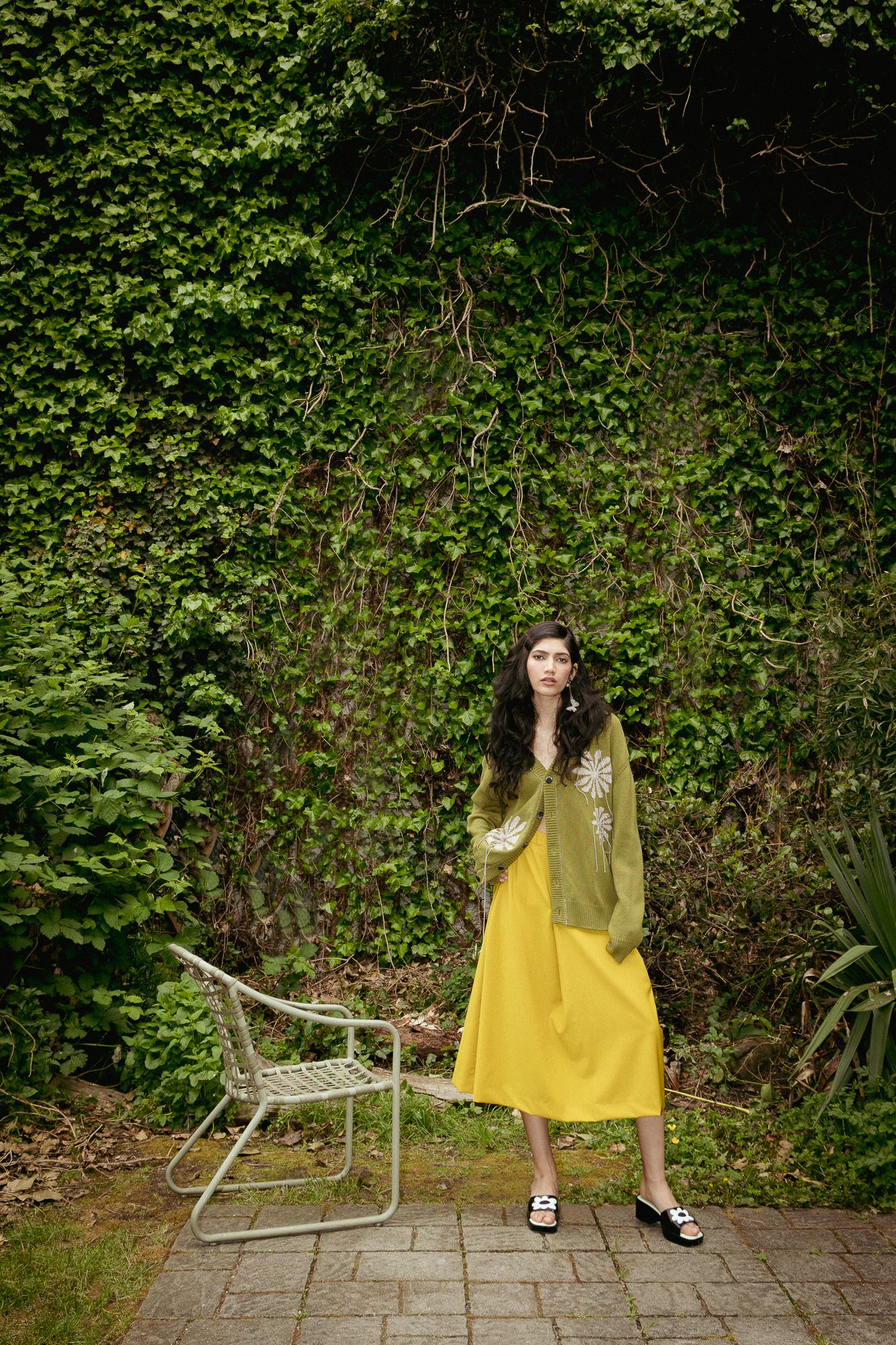 A fashion portrait of a woman in a green crochet top and yellow skirt. She's next to a chair and a vine covered wall.