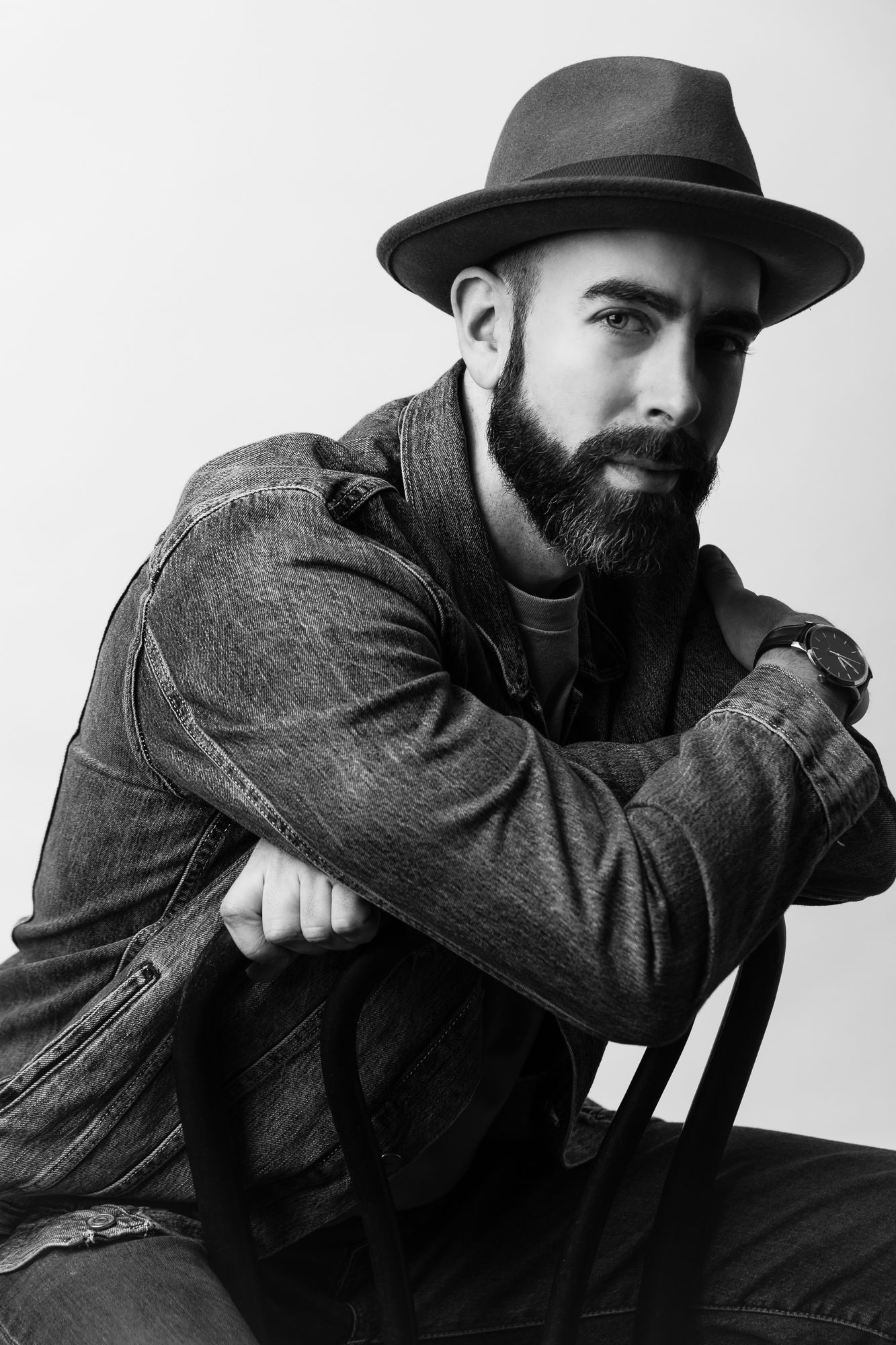 A studio headshot of a bearded man wearing a hat. He leans against the back of a chair. The lighting is dramatic.