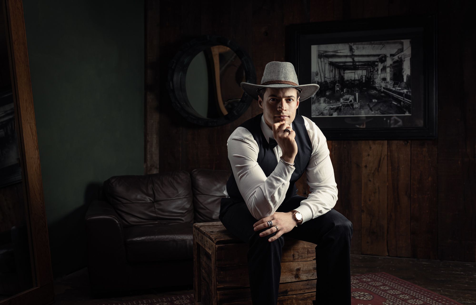 A man wears a cowboy style hat while in a tux. He has a bowtie, a nice watch, and many rings on his fingers. He sits.
