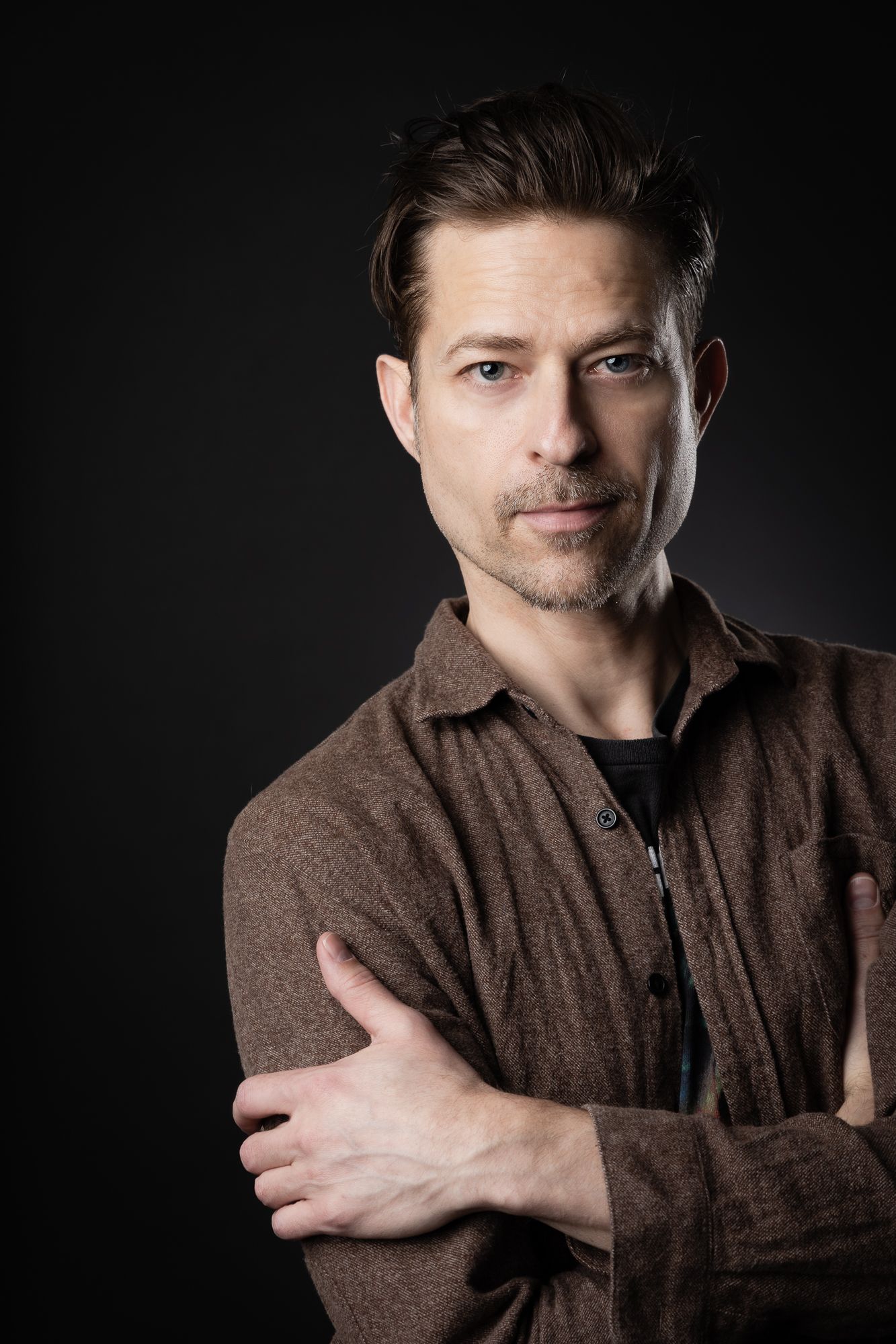 A handsome older man poses for his headshot in studio. The dramatic lighting suits him greatly. He crosses his arms.