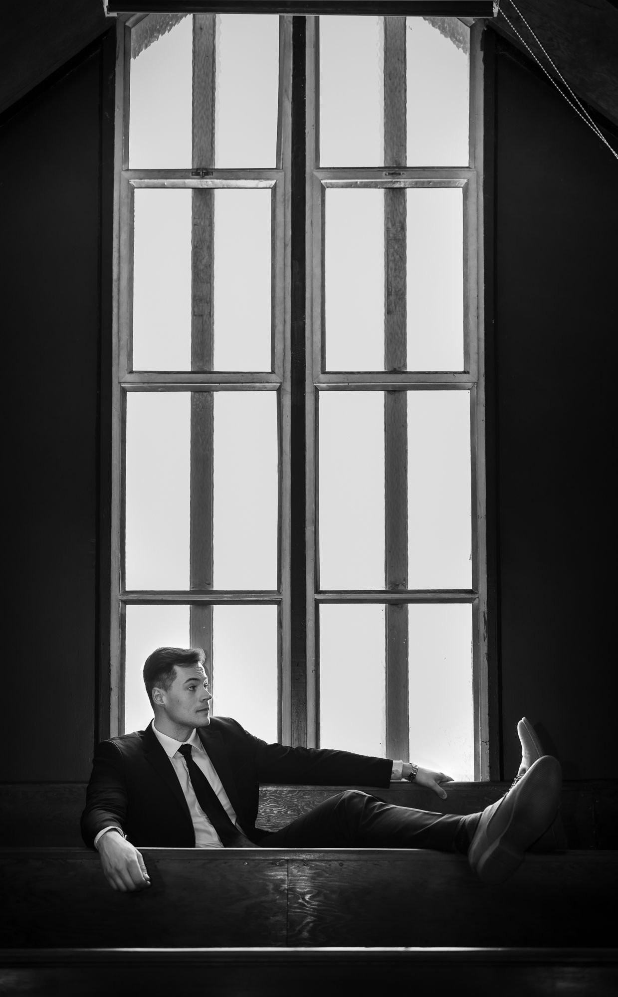 A vintage style wide-shot portrait of a handsome man in a suit leaning against a window. He looks off to the side.