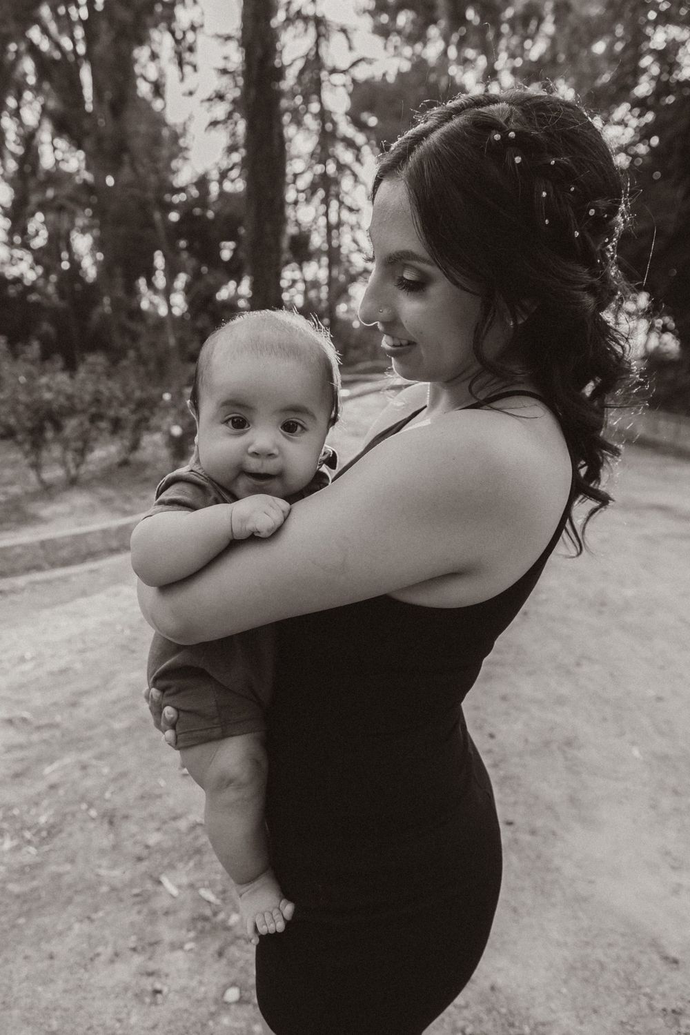 Intimate black and white photo of mother cradling infant against forest backdrop.