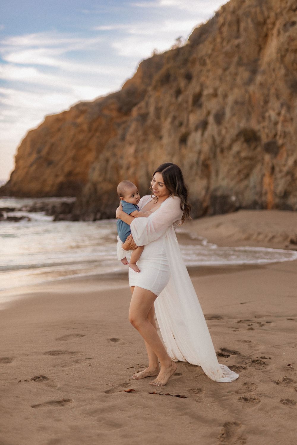 Candid beach portrait with parent holding baby against dramatic coastal cliffs.