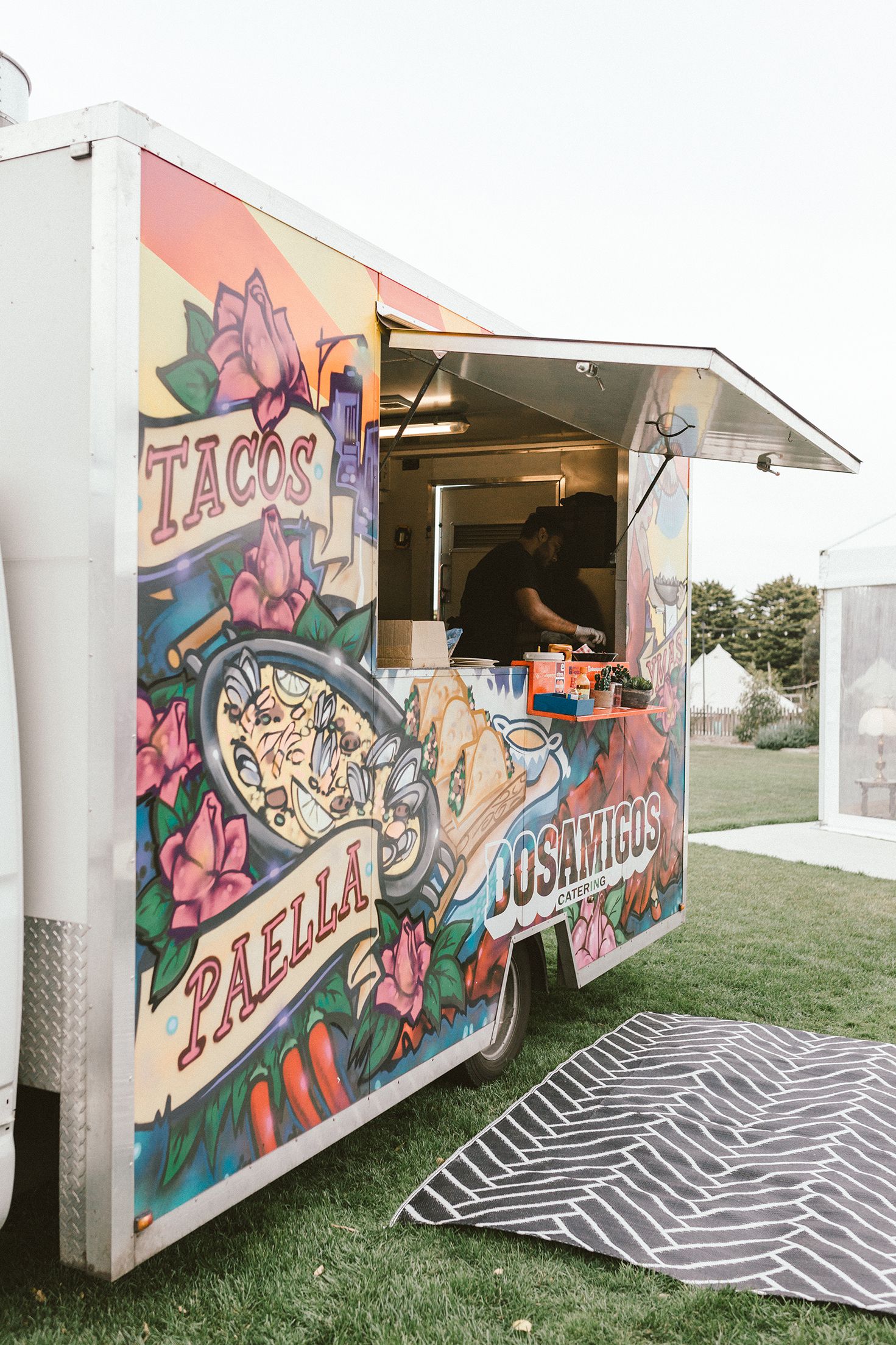 Colorful food truck with vibrant floral murals and taco signage parked on grass.