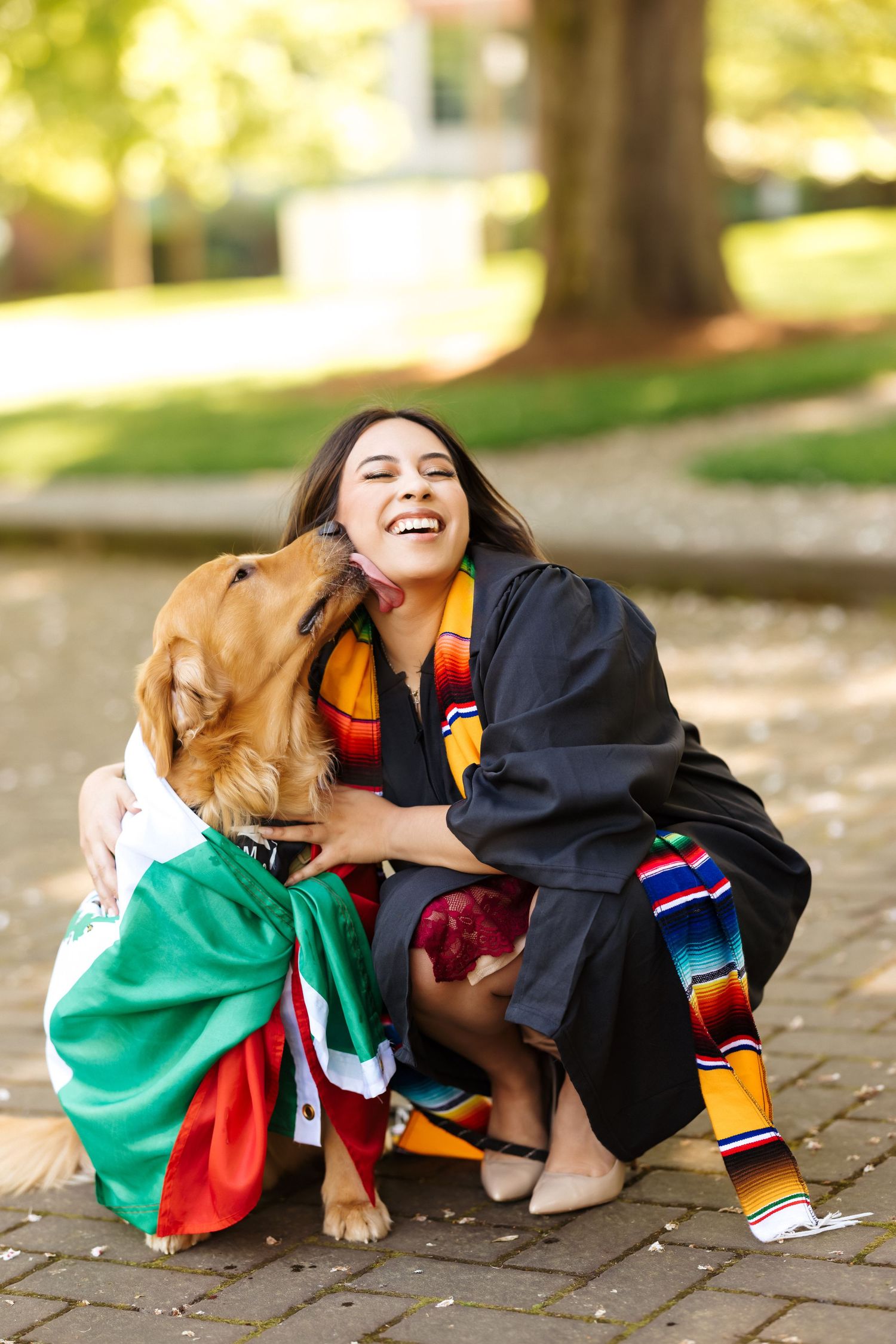 Graduate in academic regalia shares joyful moment with golden retriever wearing Mexican flag.