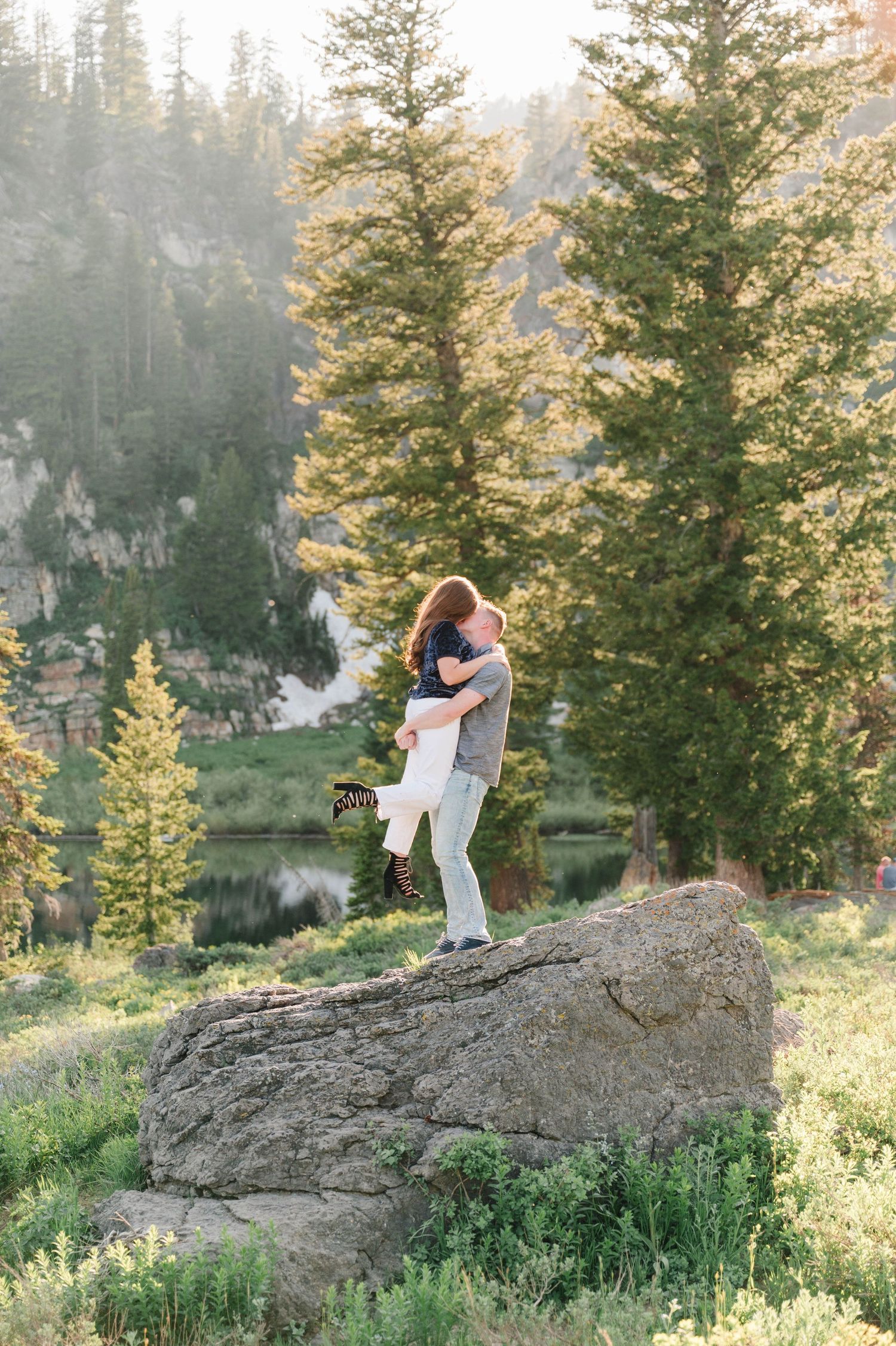 Couple embracing on large boulder with scenic forest and mountain backdrop.