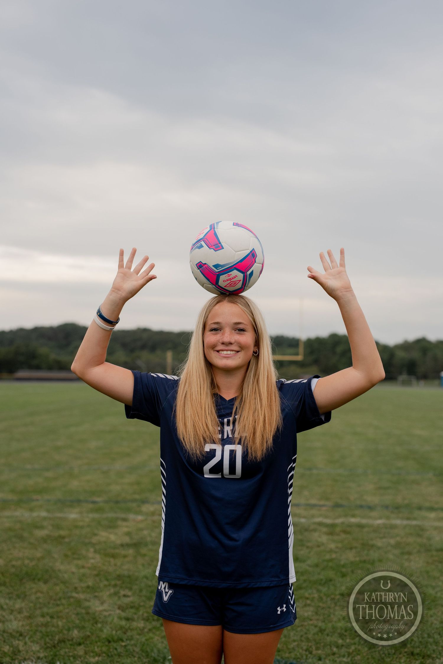 Soccer player balances colorful ball on head while raising arms on sports field.
