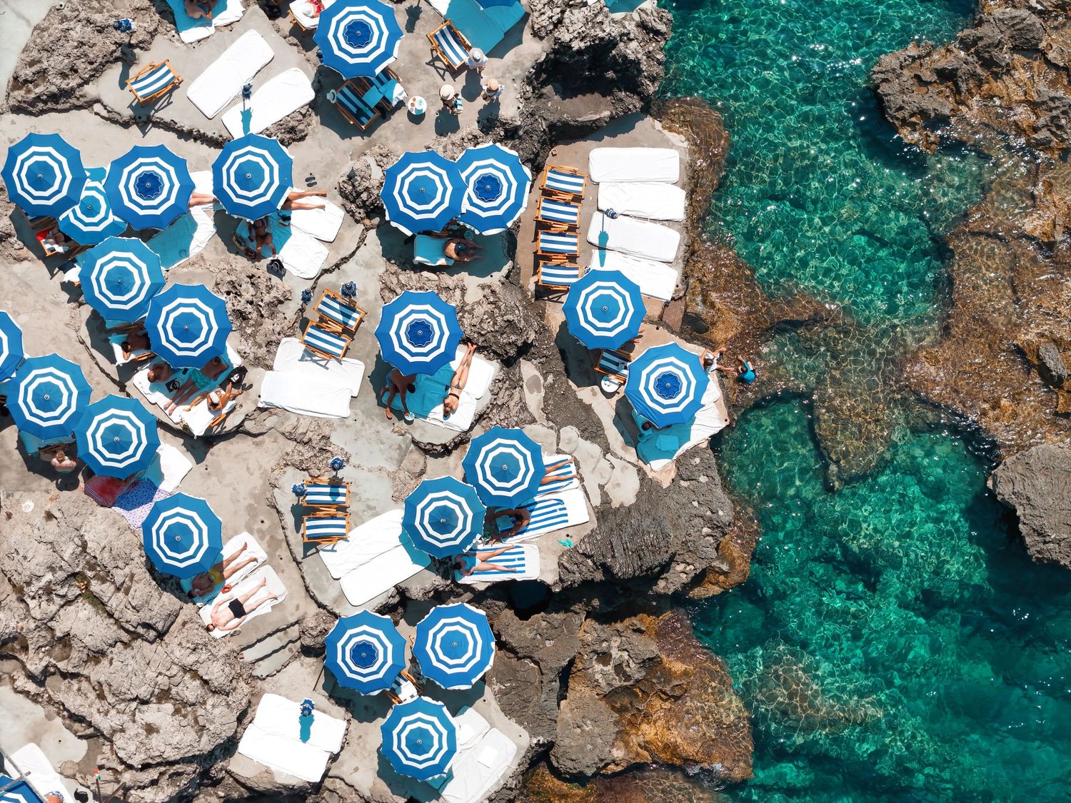 Aerial view of beach umbrellas and loungers arranged along rocky Mediterranean coastline.