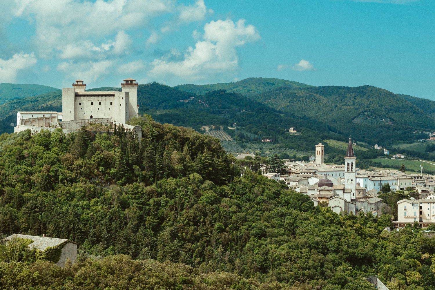 Ancient hilltop fortress overlooking a scenic Italian town surrounded by green mountains.