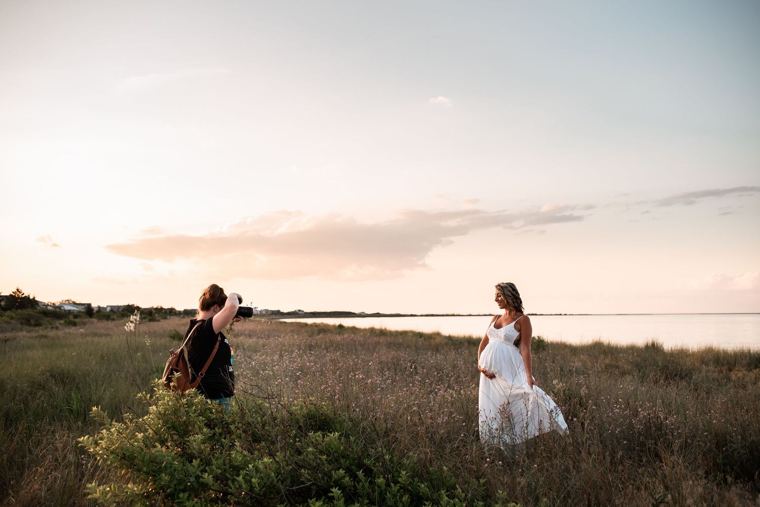 Photographer capturing a pregnant woman in a white dress standing in a golden field at sunset.