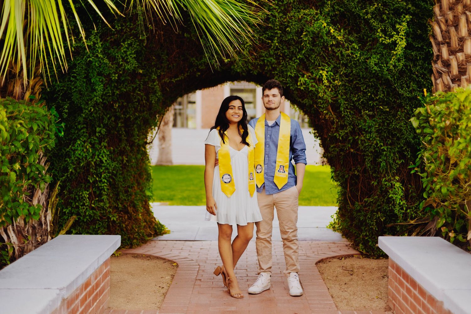 A couple in yellow and white outfits poses under an arched garden pathway.