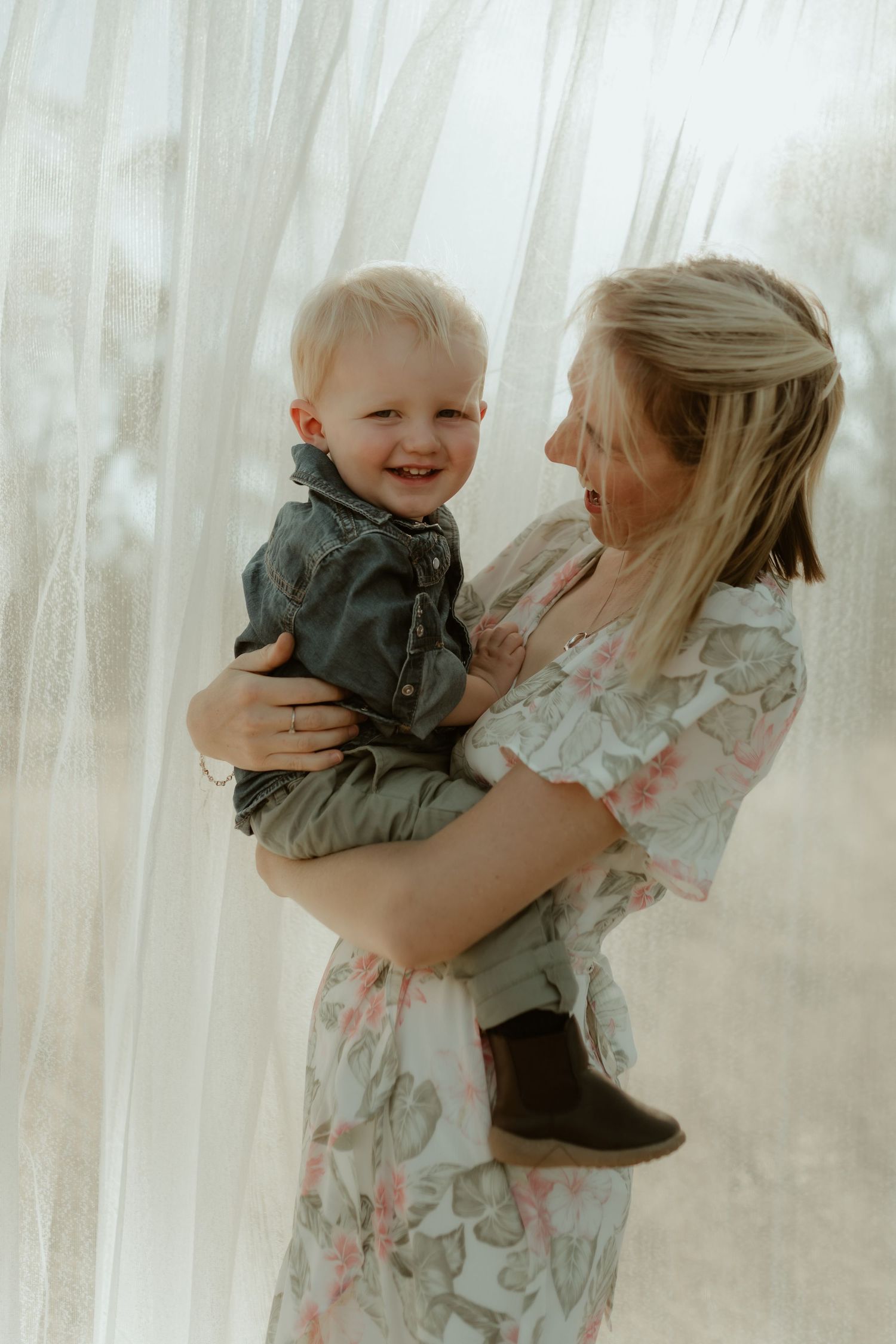 Mother in floral dress holds smiling toddler against white curtained window.
