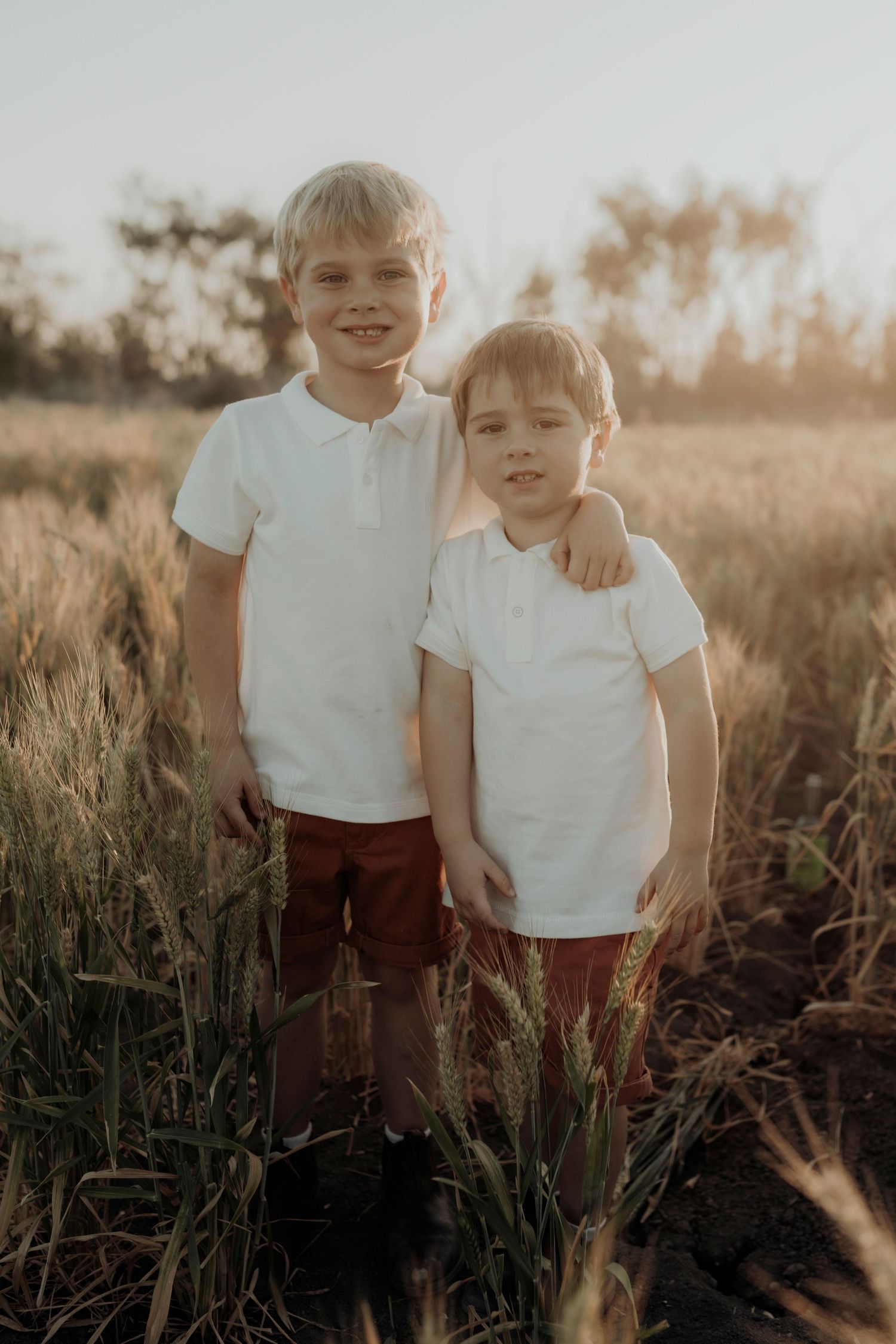 Two children in white polo shirts stand together in a sunlit meadow at golden hour.