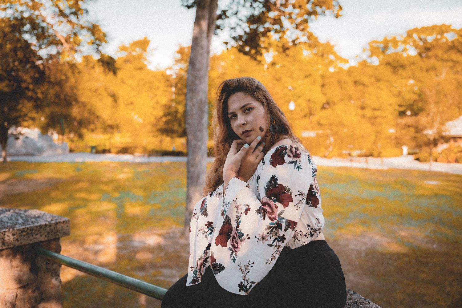 Beautiful woman sits in a white floral blouse against an autumn backdrop with golden foliage in a park setting.