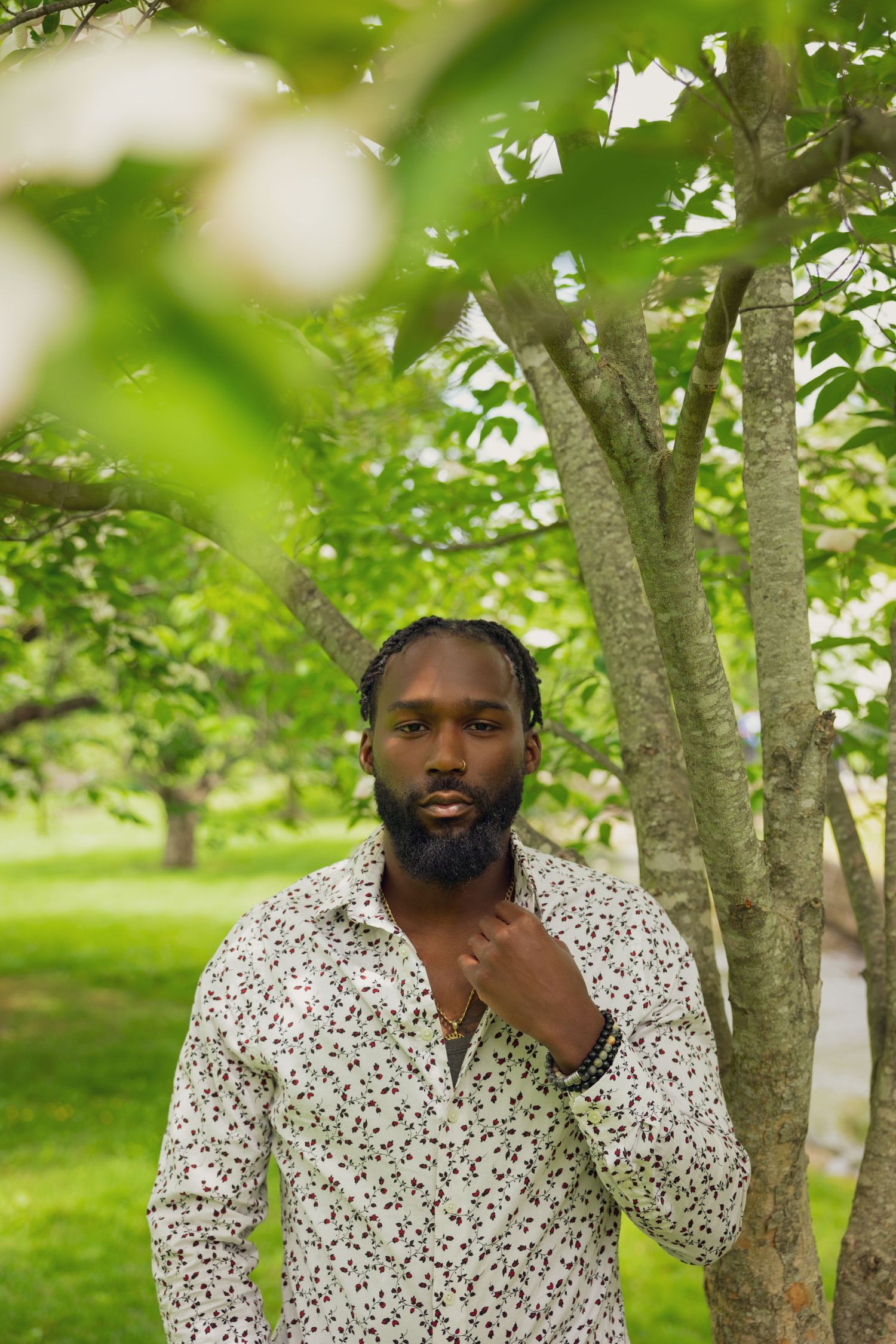 Male model wearing a floral print shirt stands against a green leafy background in a peaceful outdoor setting.