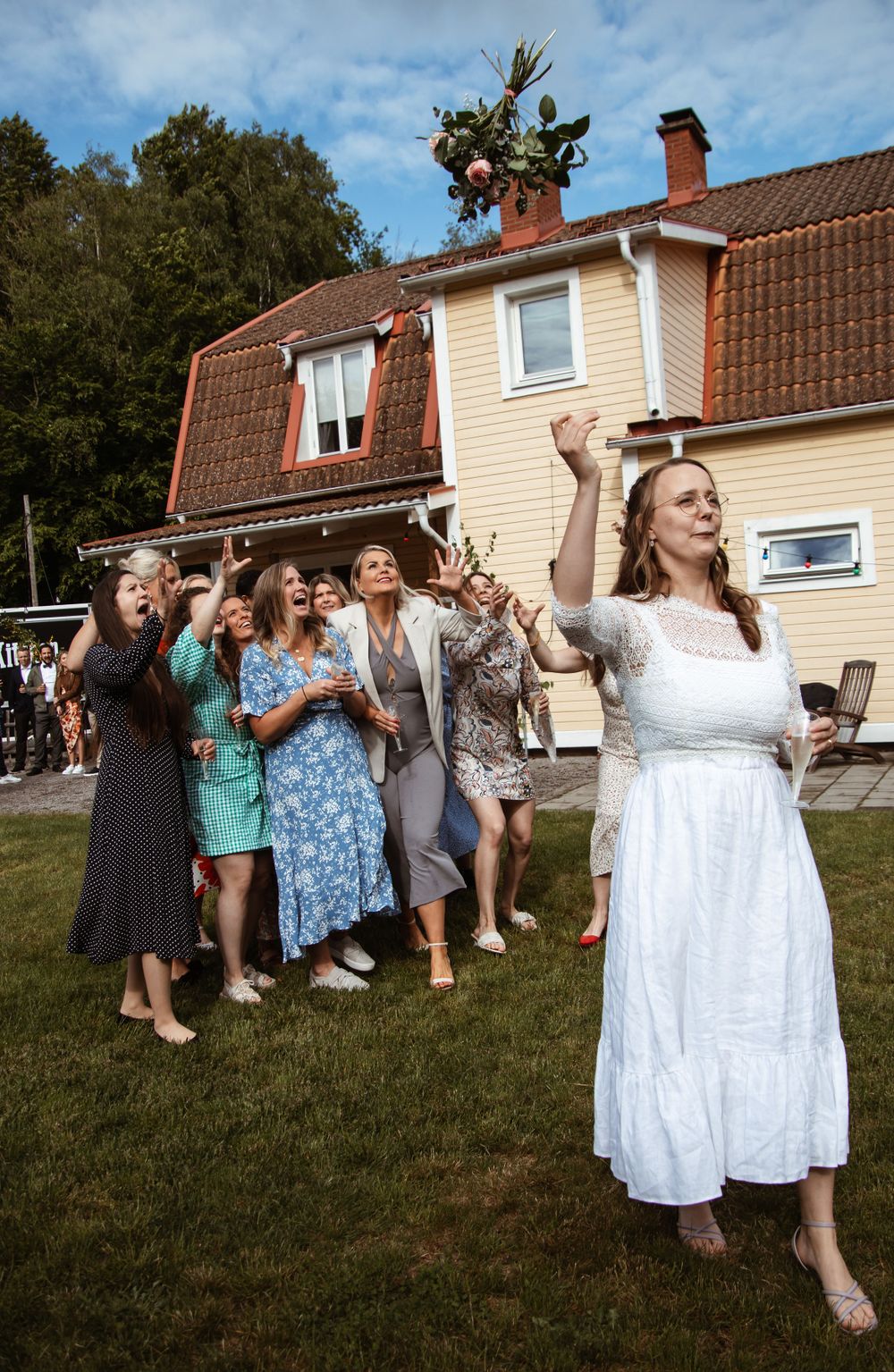 A group of people celebrates outdoors in front of a cream-colored house with a brown roof.