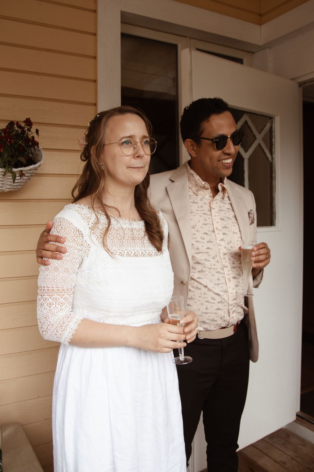 A couple in wedding attire poses together on a porch with champagne glasses.