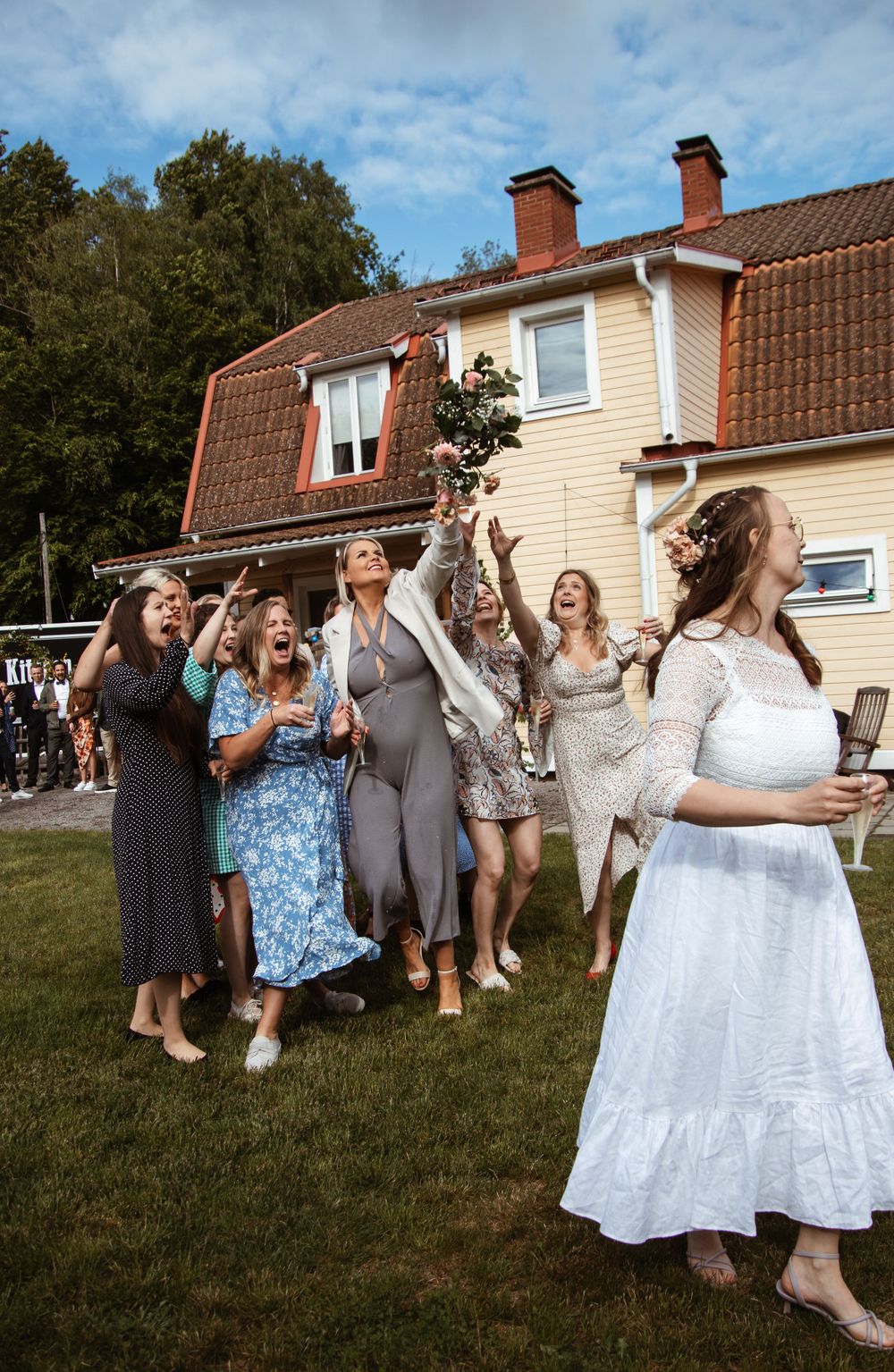 Wedding guests toss flower petals in celebration outside a house during a summer day.